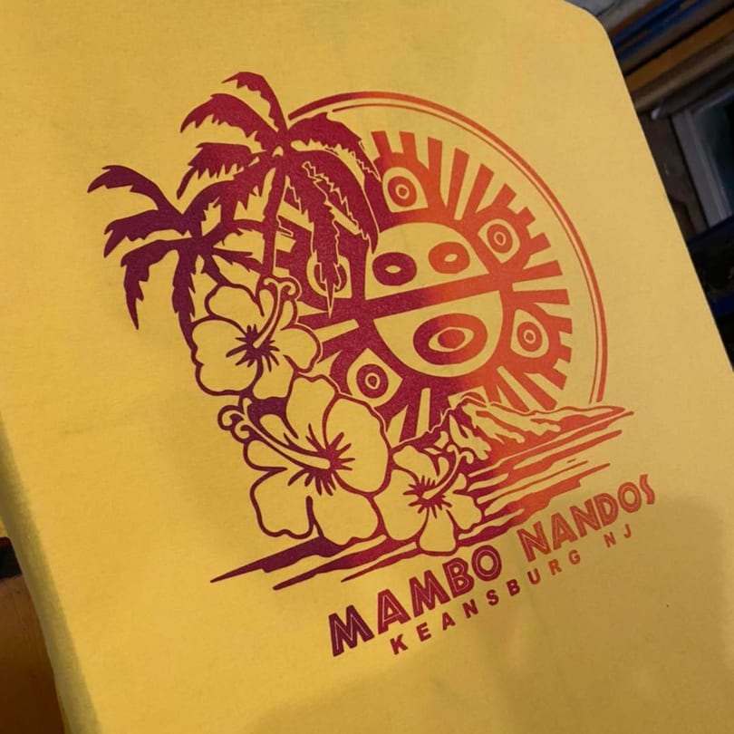 Mambo Nando's yellow t-shirt with a red and orange tropical design featuring palm trees and flowers.
