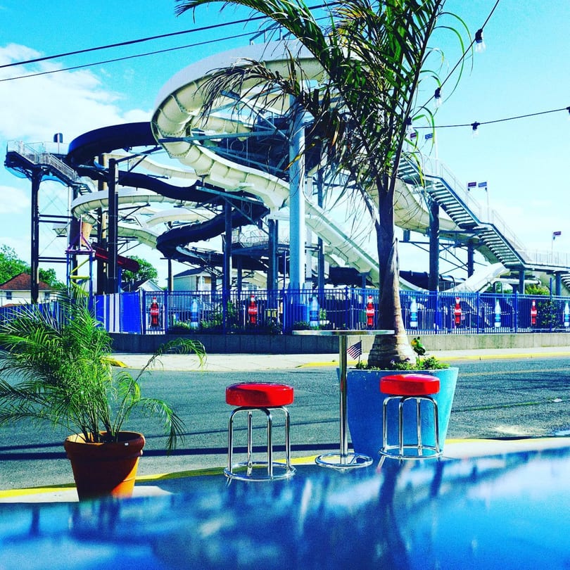 Outdoor seating with red stools and palm trees, overlooking a water park with slides.